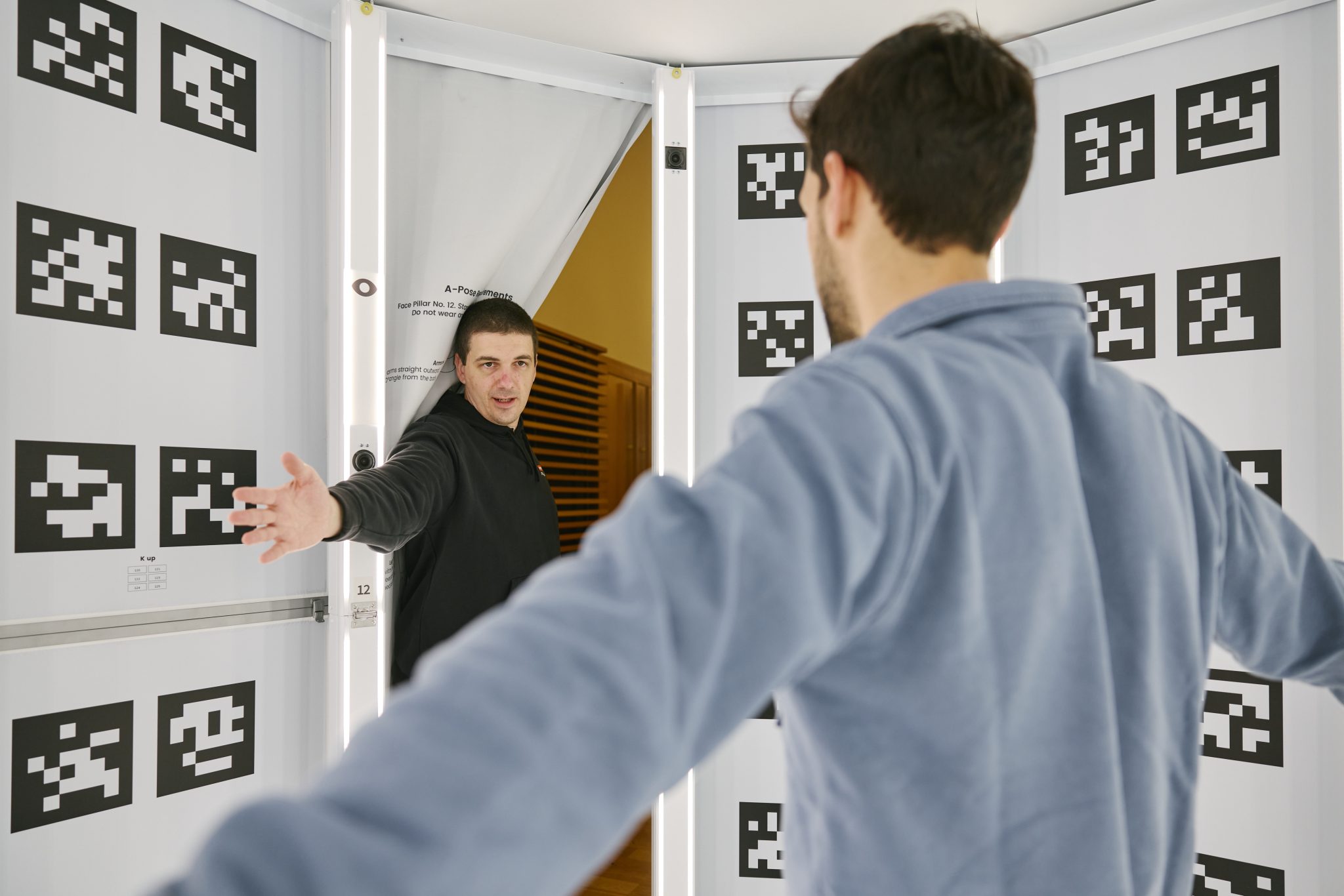 A player holds an A-pose with arms outstretched inside a body-scan booth whose walls are lined with black-and-white fiducial markers, while a technician guides from the opposite side