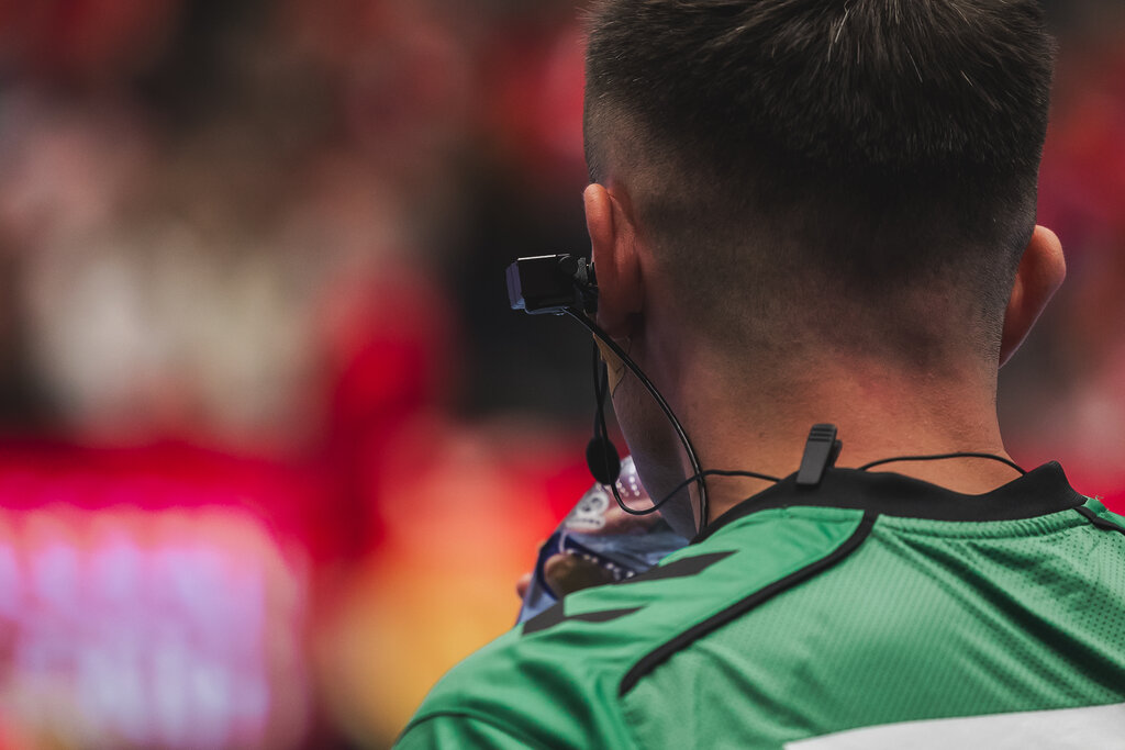 Close-up of the Riedel RefCam head unit, a small black camera worn on the side of a referee's head, with thin wires running down to the collar