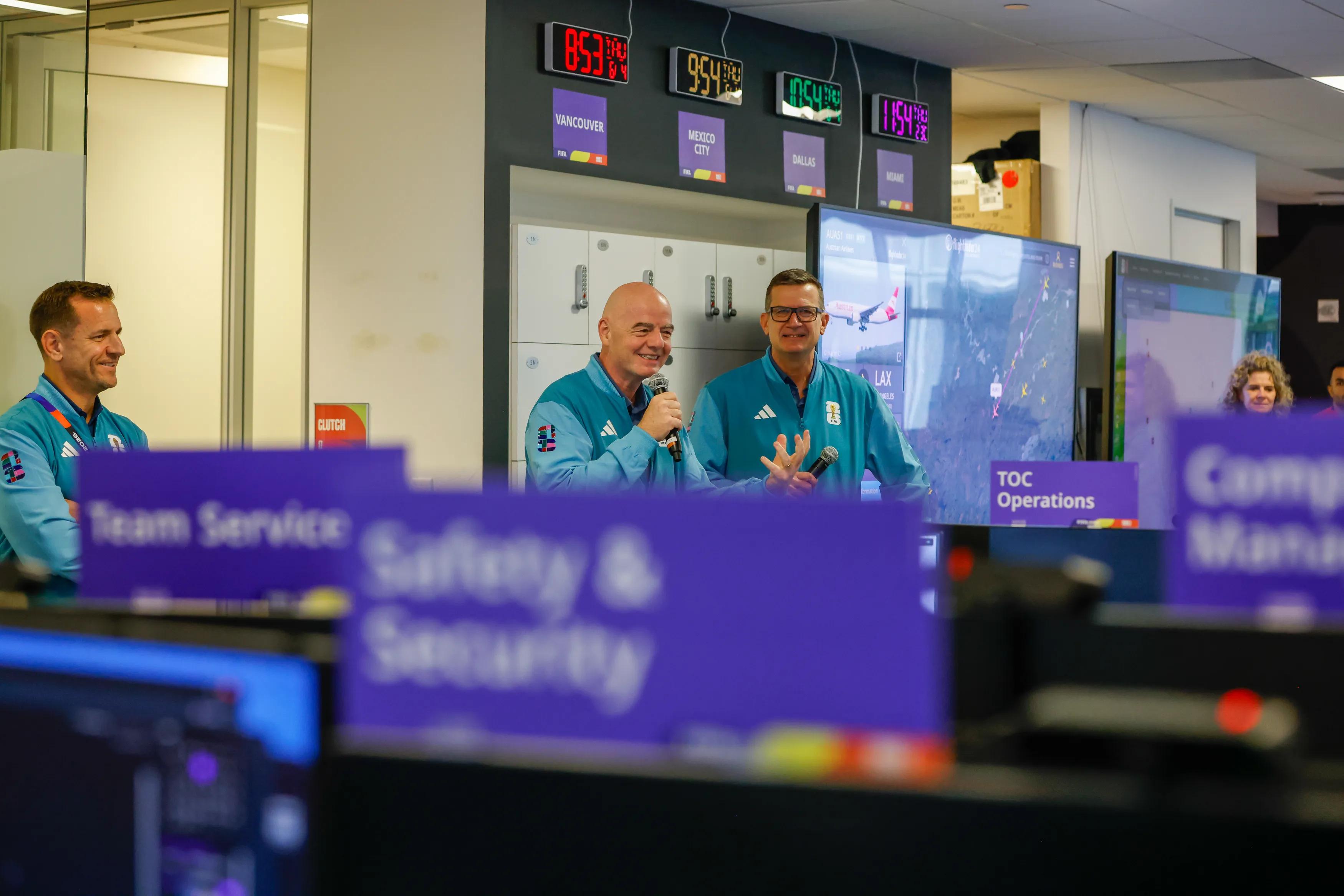 Inside FIFA's Tournament Operations Center in Miami: an executive and the FIFA President speak at a desk with a microphone in front of a wall of venue countdown clocks labeled Vancouver, Mexico City, Dallas and Miami, with large operations screens behind and desk panels reading Team Service, Safety and Security, Competition Management and TOC Operations